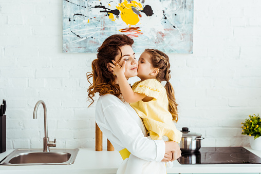 cute daughter kissing smiling beautiful mother in kitchen