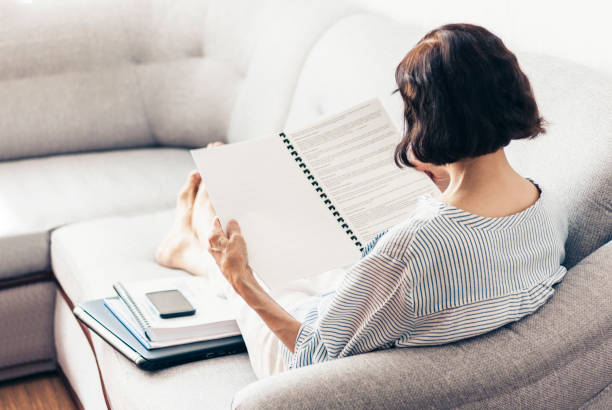 istock Middle-aged brunette woman on the gray sofa reading documents, using laptop and smartphone 1152885722