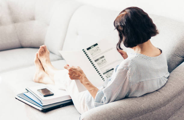 istock Middle-aged brunette woman on the gray sofa reading documents, using laptop and smartphone 1152886018