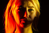 Close up portrait of face an serious girl with bright green eye looking into camera: straight nose, expressive eyes, ears, blonde hair illuminated with orange, yellow and red light on black background