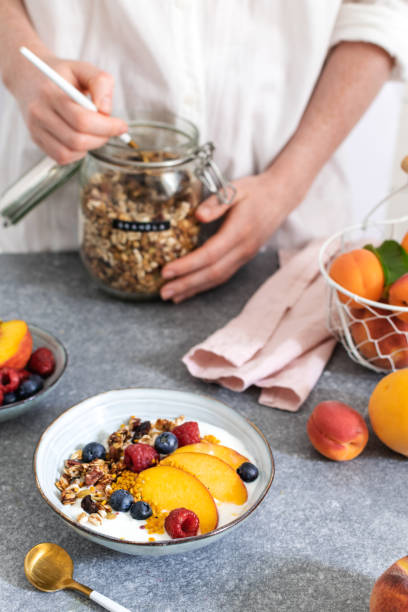 istock Female hands serving granola from jar, with greek yogurt fresh peach, blueberry, raspberry on the table - healthy breakfast 1152913276