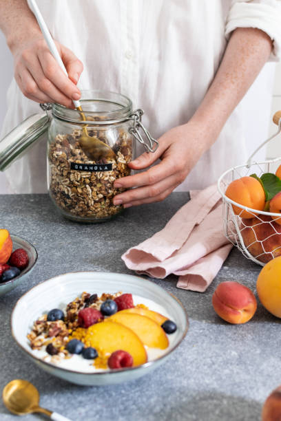 istock Female hands serving granola from jar, with greek yogurt fresh peach, blueberry, raspberry on the table - healthy breakfast 1152913284
