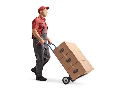 Full length shot of a young male worker in a uniform pushing boxes on a hand truck isolated on white background