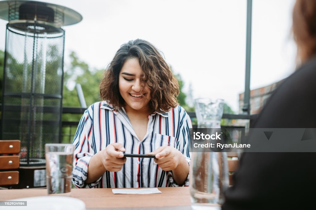 Hispanic Woman Photographing Check for Mobile Bank Deposit A woman out to lunch with friends photographs her paycheck with her smartphone to deposit it in her bank account. Banking Stock Photo