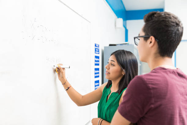 She's Almost Got It Teenage girl solving equation on whiteboard while standing with classmate in school doing a favor stock pictures, royalty-free photos & images