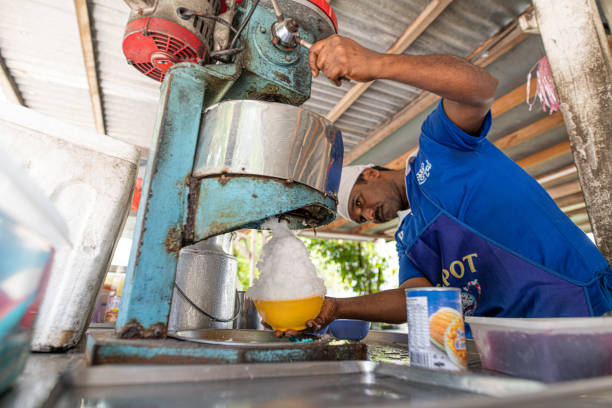 un venditore ambulante indiano che prepara "ice kacang" un tradizionale deserto dolce con ghiaccio e zucchero di canna, arachidi, fagioli rossi e latte nella sua bancarella nel quartiere del centro di melaka - syrup brown sugar sugar spoon foto e immagini stock