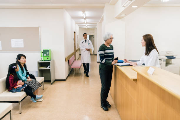 Senior woman talking to receptionist in hospital waiting area Japanese woman standing in waiting room, talking to staff, assistance, support, hospital interior doctor waiting room stock pictures, royalty-free photos & images