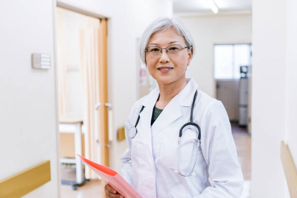 Portrait of attractive senior female doctor holding notes Japanese doctor with short grey hair and glasses, smiling towards camera in hospital corridor, confidence, efficiency, pride japanese female doctor stock pictures, royalty-free photos & images