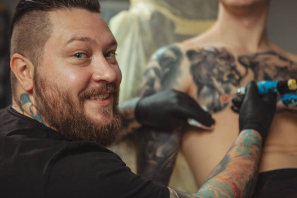 Man getting new tattoo by professional tattooist Close up of a cheerful bearded tattoo artist smiling to the camera over his shoulder, while tattooing his client on chest. Happy professional tattooist working at his studio. Creativity concept chest tattoo pain stock pictures, royalty-free photos & images