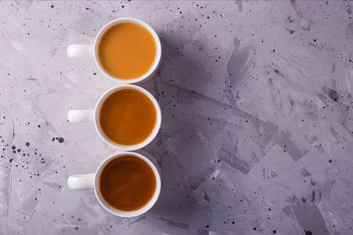 Three cups of masala tea or coffee with milk of different colors on a gray textural background. Top view, flat lay. Copy space