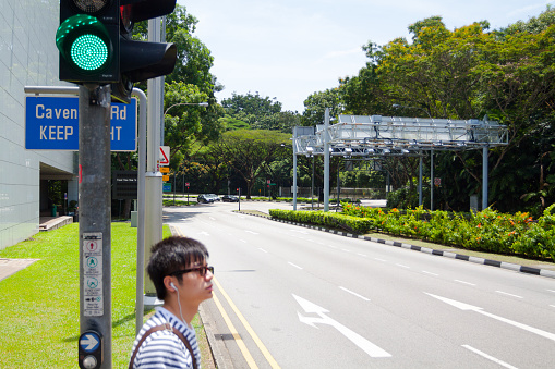 Young asian man waiting at traffic light on street in university area in Singapore Queenstown in western part of country. Above man is green light for traffic. In background are trees.