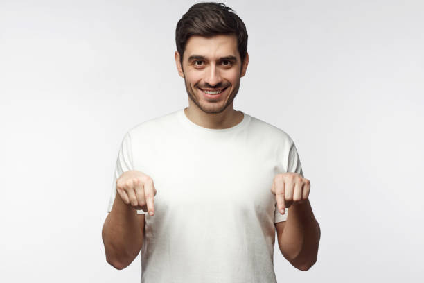 привлекательный молодой человек в белой футболке, указывая двумя пальцами, изолированный на сером фоне - male fashion model adult human arm стоковые фото и изображения