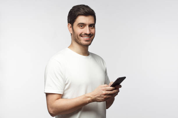 istock Young man looking and smiling at the camera with cheerful expression while texting to friend, using mobile phone, isolated on gray background 1153003761