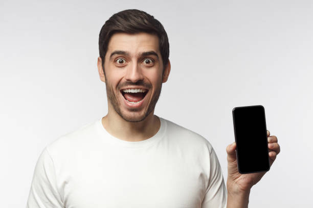 istock Excited young man holding blank phone with empty black screen, screaming WOW, isolated on gray background 1153003768