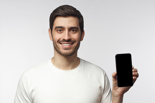 Young smiling man showing blank screen of smart phone in hand, isolated on gray background