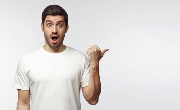 istock Young surprised man in white t-shirt looking at camera with open mouth, pointing right, copy space for ads, isolated on gray background 1153003773
