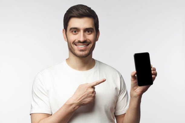 istock Young smiling man holding blank phone with empty black screen, pionting to display with finger, isolated on gray background 1153003785