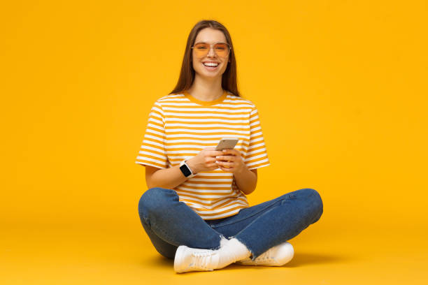 bella donna sorridente seduta sul pavimento con le gambe incrociate e con in mano il cellulare, isolata su sfondo giallo - floor sitting foto e immagini stock