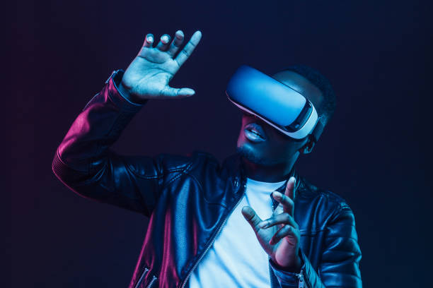 istock Young african man wearing virtual reality goggles with hands up, isolated on black background 1153003868