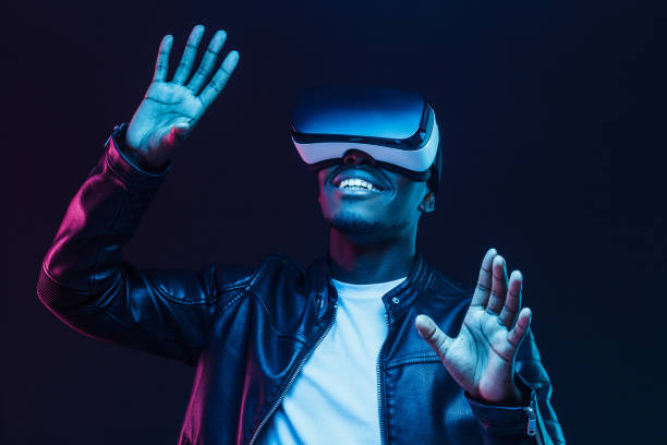 istock Young african man wearing virtual reality goggles with hands up, isolated on black background 1153003884