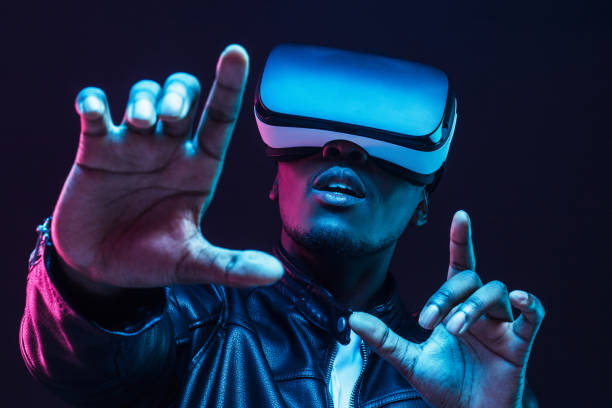 istock Young african man wearing virtual reality goggles with hands up, isolated on black background 1153003888