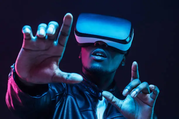 Photo of Young african man wearing virtual reality goggles with hands up, isolated on black background