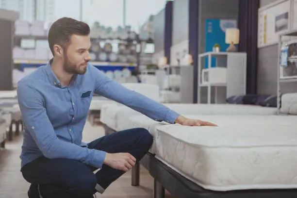 Young handsome man examining orthopedic mattress on sale at furniture store, copy space. Attractive male customer buying new bed at homeware supermarket. Consumerism, home concept