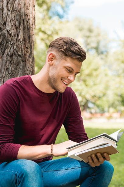 istock cheerful student reading book and smiling while sitting in park 1153041281