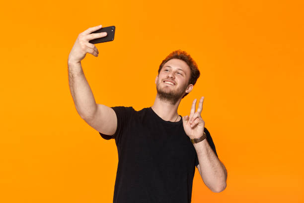 istock Studio shot of a smiling bearded young man making selfie with the smartphone while standing over orange background 1153042857