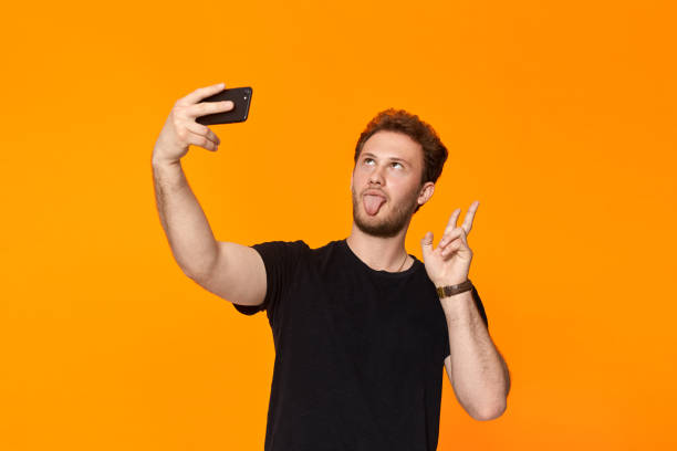 istock Studio shot of a smiling bearded young man making selfie with the smartphone while standing over orange background 1153042858