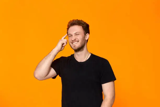 Studio shot of a bearded young man pointing a finger at the temple while standing over orange background. Concept of a positive thinking.