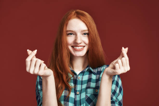 Speading love and happiness. Redhead girl showing heart sign Portrait of a beautiful caucasian girl in checkered shirt smiling and showing mini heart sign hands. Isolated on red background. Korean mini heart finger sign. Asian symbol hand heart. Korean, Chinese and Japanese Symbol hand heart little redhead girl stock pictures, royalty-free photos & images