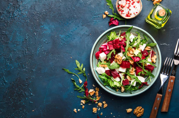 istock Beet summer salad with arugula, radicchio, soft cheese and walnuts on plate with fork, dressing and spices on blue kitchen table, copy space, top view 1153071435