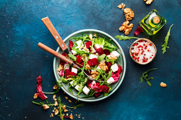 istock Beet summer salad with arugula, radicchio, soft cheese and walnuts on plate with fork, dressing and spices on blue kitchen table, copy space, top view 1153071465