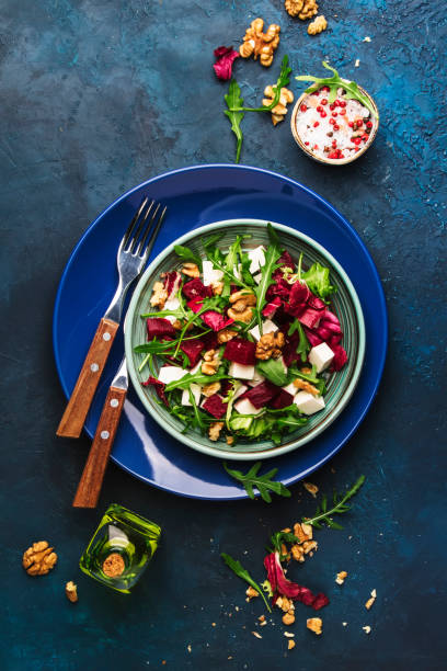 istock Beet summer salad with arugula, radicchio, soft cheese and walnuts on plate with fork, dressing and spices on blue kitchen table, copy space, top view 1153071513