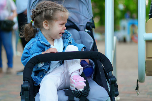 Little Caucasian girl dressed in a denim jacket sits in a children's carriage and cries. Children's whims and tantrums