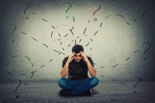 istock Confused and tired young man seated on the floor keeps hands to forehead looking down thoughtful surrounded by different tasks to solve as having many questions. 1153091015