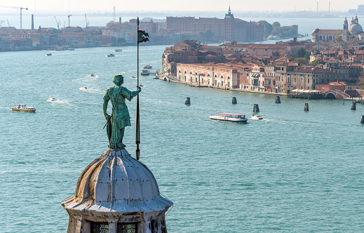 An aerial overview of a bronze statue, standing at the top of the dome of Basilica of San Giorgio Maggiore, watching over the busy Giudecca Canal. Venice, Veneto, Italy.