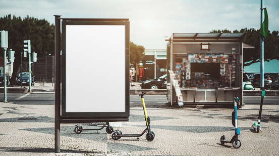 Mockup of an empty urban advert poster surrounded by scooters scattered across the street on the pavement stone; template of a blank street information billboard with a newsstand in the background
