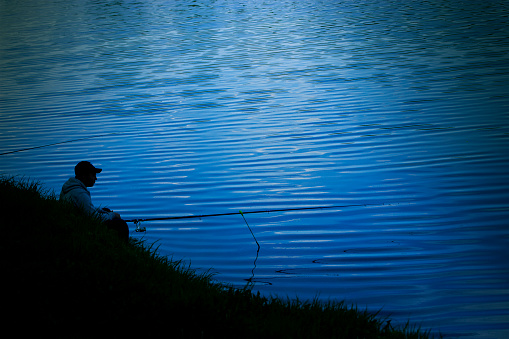 Night fishing, dark silhouette of a fisherman with a fishing rod sits on the shore the background of dark blue water, a man catches fish in the moonlight