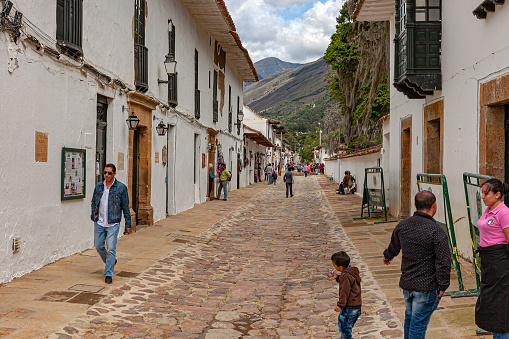 Villa de Leyva, Colombia - November 26, 2017: looking up Carrera 9 from Calle 13 on the Plaza Mayor in the historic 16th Century colonial town of Villa de Leyva in the Boyacá Department. It is noon and some local people are seen on the Carrera; in the far background is a section of the Andes Mountains. Founded in 1572 and located at just over 7000 feet above mean sea level on the Andes Mountains, Villa de Leyva was declared a National Monument by the Colombian Government in 1954 to protect its colonial architecture and heritage. The Town was also one of the locations for the movie Cobra Verde by Werner Herzog, and the Spanish language Soap Opera Zorro. Photo shot in the midday sunlight; horizontal format. Copy space.