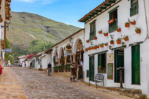 Villa de Leyva, Colombia - November 26, 2017: looking up Calle 13 in the historic 16th Century colonial town of Villa de Leyva in the Boyacá Department. To the right are a row of restaurants one after the other, with a member of staff standing on the street trying to entice customers into their restaurants. In the far background is a section of the Andes Mountains. Founded in 1572 and located at just over 7000 feet above mean sea level on the Andes Mountains, Villa de Leyva was declared a National Monument by the Colombian Government in 1954 to protect its colonial architecture and heritage. The Town was also one of the locations for the movie Cobra Verde by Werner Herzog, and the Spanish language Soap Opera Zorro. Photo shot in the midday sunlight; horizontal format. Copy space.