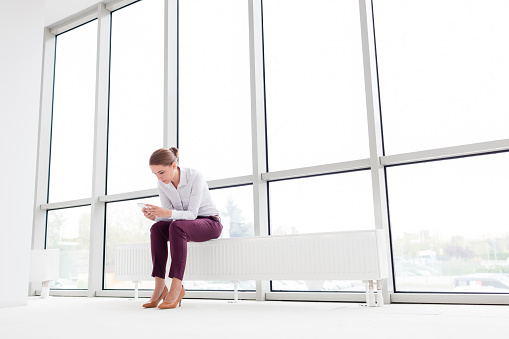 Young businesswoman using smartphone while sitting on radiator at new empty office