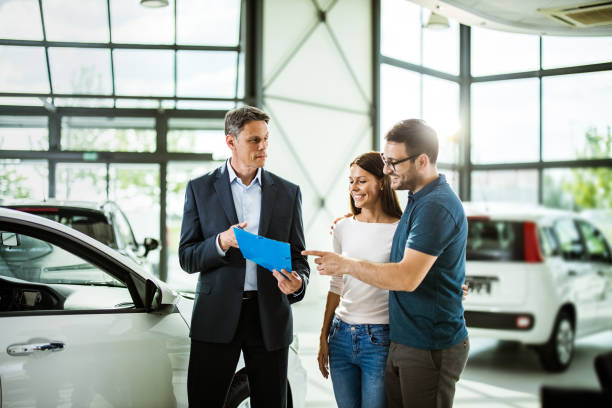 Happy embraced couple and car salesperson going through paperwork in a showroom. Happy couple reading buying plan with car salesperson in a showroom. car ownership photos stock pictures, royalty-free photos & images