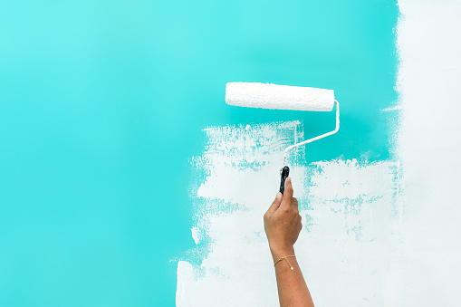 Woman painting over turquoise colored wall with paint roller.