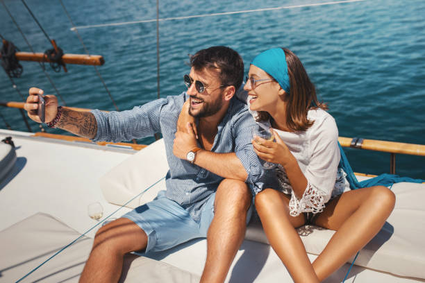 Young couple taking selfies on a sailing cruise. Closeup front angle view of a mid 20's couple enjoying their sailing cruise. They are sitting on the deck and snapping some selfies to make memories and of course to boost their social feed. yacht selfie stock pictures, royalty-free photos & images