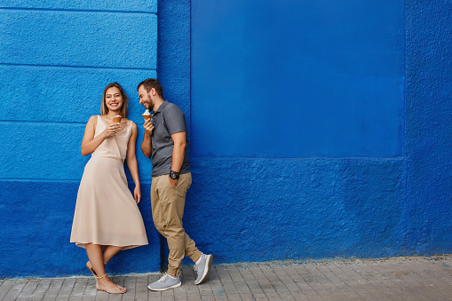 Happy couple having fun, eating ice cream and smiling against the blue wall