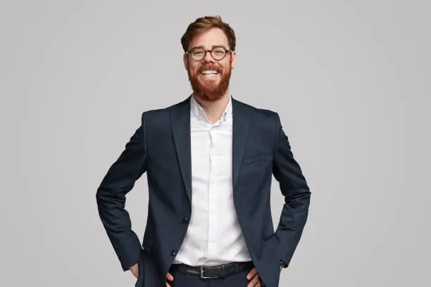 Successful Irish businessman with ginger beard cheerfully smiling and looking at camera while standing against gray background