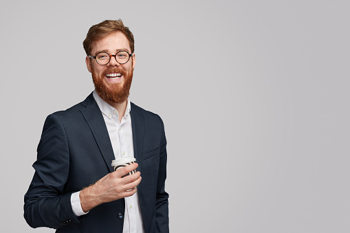 Attractive ginger bearded male with cup of fresh hot drink smiling and looking at camera while standing against gray background during break