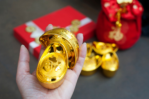 Chinese gold ingots on hand with Chinese character meaning whealth, rich, gold, luck, healthy. Gift box and red pouch as a background. Chinese New Year Gift concept.